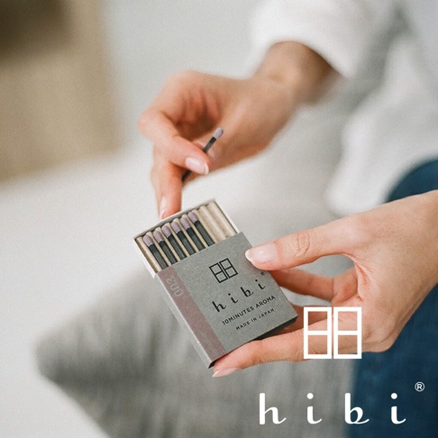 一雙手輕輕拿著一個灰色日系品牌 hibi 的小型火柴盒，盒內整齊排列著數根帶有紫色火柴頭的香氛火柴，其中一根火柴已被取出。包裝盒印有 hibi 品牌標誌與 10minutes aroma 字樣，整體氛圍簡約質感，呈現出日式精緻的生活美學。