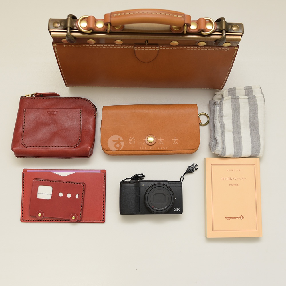 職人手作皮革復古口金兩用手提包，搭配紅棕色拉鍊皮夾、焦糖色長夾、卡夾、黑色的數位相機、條紋布料和黃色書籍。