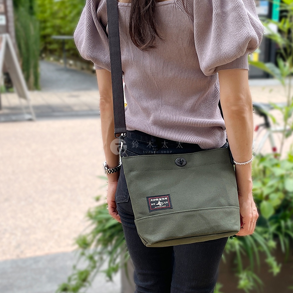 軍綠色純棉帆布迷你側背包，正面有一塊帶有日文圖案的貼布和一個黑色按扣。背包透過黑色斜揹帶固定在肩上。