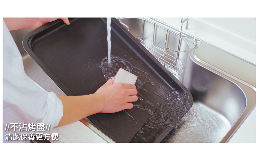 左手正在用白色海綿清潔黑色電烤盤，水流從烤盤一側傾倒至水槽，烤盤邊緣有圓潤的設計。