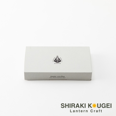 淺灰色紙盒，盒蓋中央印有黑色達摩不倒翁圖案與日文「マメココラン」字樣，下方印有英文「mame cocolan mini-chochin from Yame, Japan」。