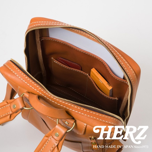 職人手作皮革軟皮商務後背包，焦糖棕色，展示打開的主隔層，內有隔袋，可放置卡片、筆記本和橘色小筆記本。包包採用對比色縫線和金色拉鍊。