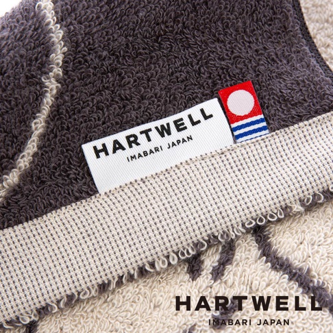 這是一張展示 Hartwell 今治毛巾細節的特寫照片，深灰色與米色的毛圈布料上縫有品牌標籤，標籤上印有 Hartwell Imabari Japan 字樣及紅白色圓形圖標，畫面右下角亦有相同的品牌文字。