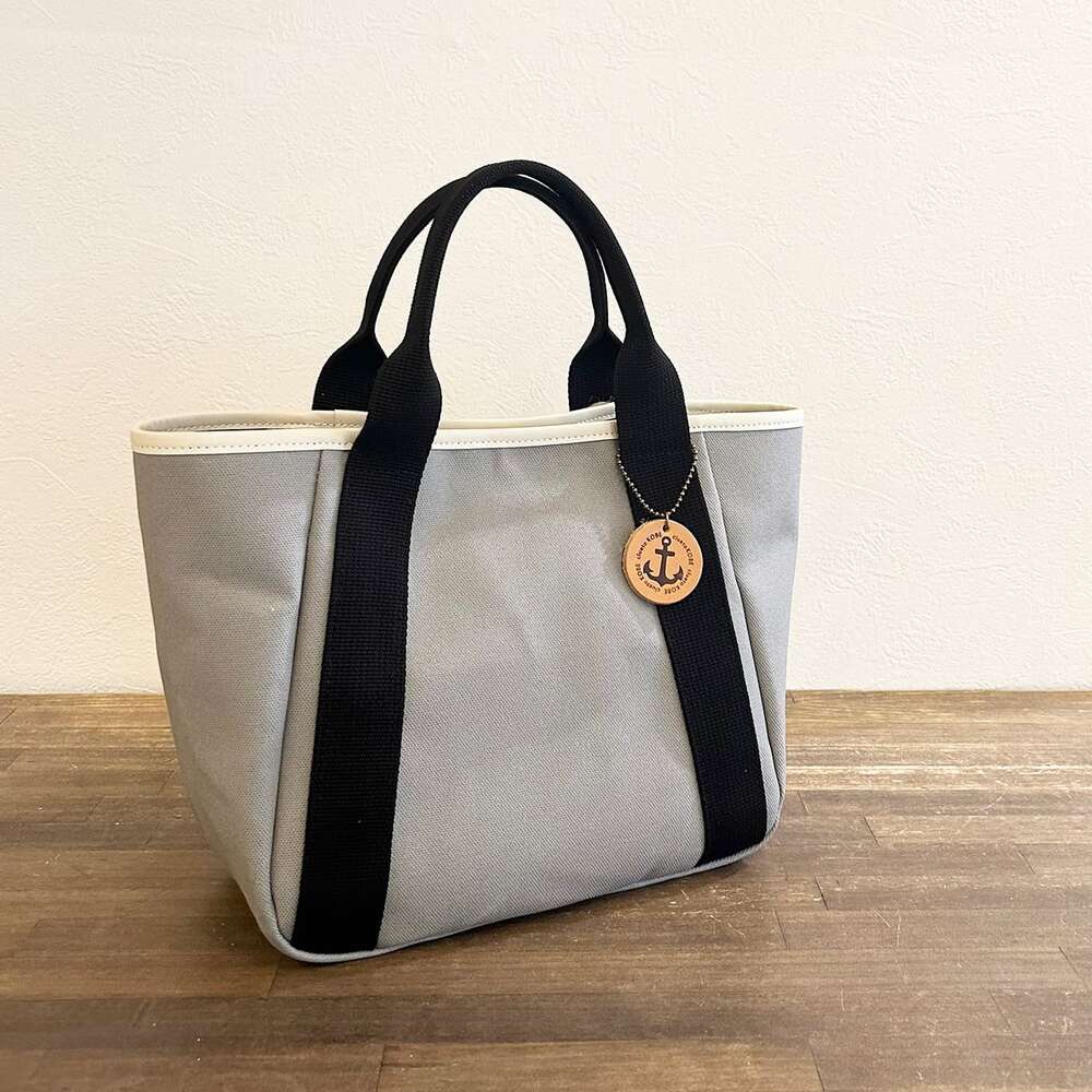 淺灰色帆布托特包，配有黑色提把和白色滾邊，包包一側掛著一個帶有船錨圖案的圓形木質吊飾。