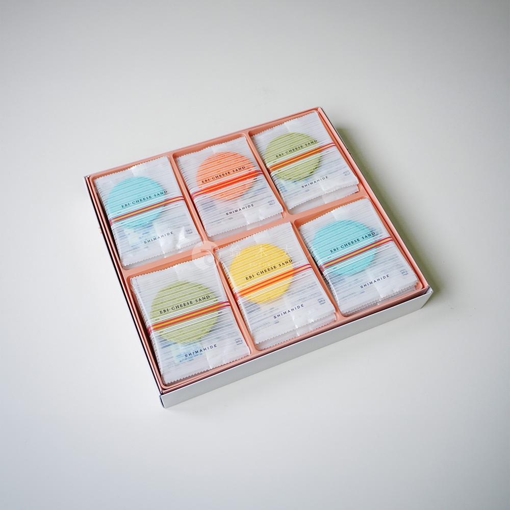 這是一個禮盒包裝的俯視視角照片，盒內整齊排列著六包獨立密封的志滿秀馬卡龍起司蝦仙貝，每包包裝紙上皆印有 EBI CHEESE SAND 及 SHIMAHIDE 字樣，內部透出藍、橘、綠、黃等多種鮮豔繽紛的色彩，展現出精緻且充滿和洋風格的日式伴手禮質感。