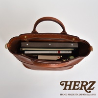 職人手作皮革大容量兩用托特包，棕色皮革材質，內部可見多本書籍、筆記本，拉鍊開合，附有提把及側邊金屬環。