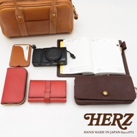 HERZ 品牌皮革製品，包含一個棕色迷你波士頓包，一個黑色相機，一本日誌本，一個紅色零錢包，一個紅色卡夾，以及一個深棕色皮夾。