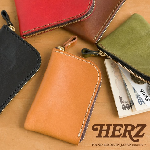 木紋桌面上擺放著五個日本HERZ職人手作皮革分隔收納迷你錢包，錢包呈現紅、棕、黑、淺棕及綠色。右側綠色錢包拉鍊開啟，內部裝有紙鈔與硬幣。右下角印有HERZ品牌名稱與標語。