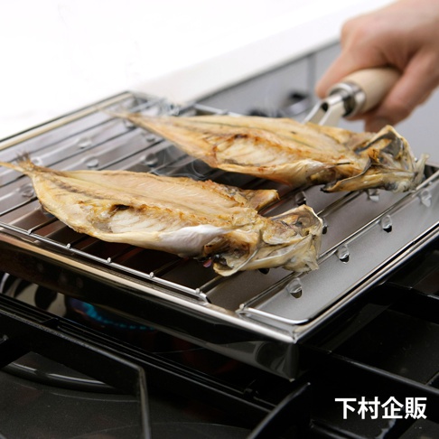 瓦斯爐專用炭烤風萬用燒烤盤，不鏽鋼材質，黑色爐架上放置著兩條烤製金黃的魚，下方有藍色火焰，右上角有一隻手持木柄的夾子。