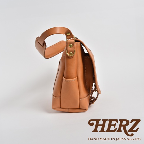 這款復古郵差包採用淺棕色皮革製成，側邊可見兩個小口袋，包身結構飽滿，搭配圓潤的提把和金屬扣環，展現職人手作質感。