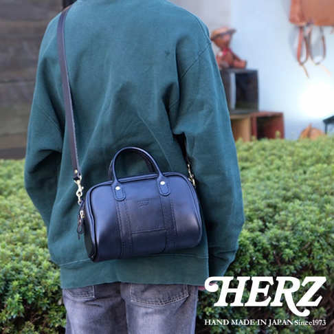 一位身穿深綠色連帽衫的男士，斜挎著一個黑色皮革迷你波士頓包，包包上有HERZ的Logo，背景是綠色植物和一間商店。