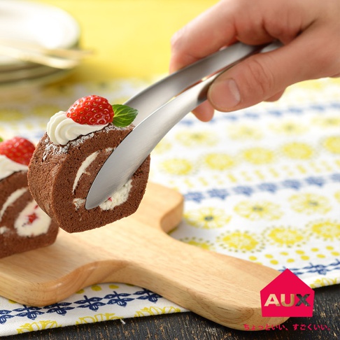 銀色金屬材質的leye 不沾桌配料專用夾，夾取著裝飾有鮮奶油與草莓的巧克力瑞士捲，夾子前端呈現流線型設計。