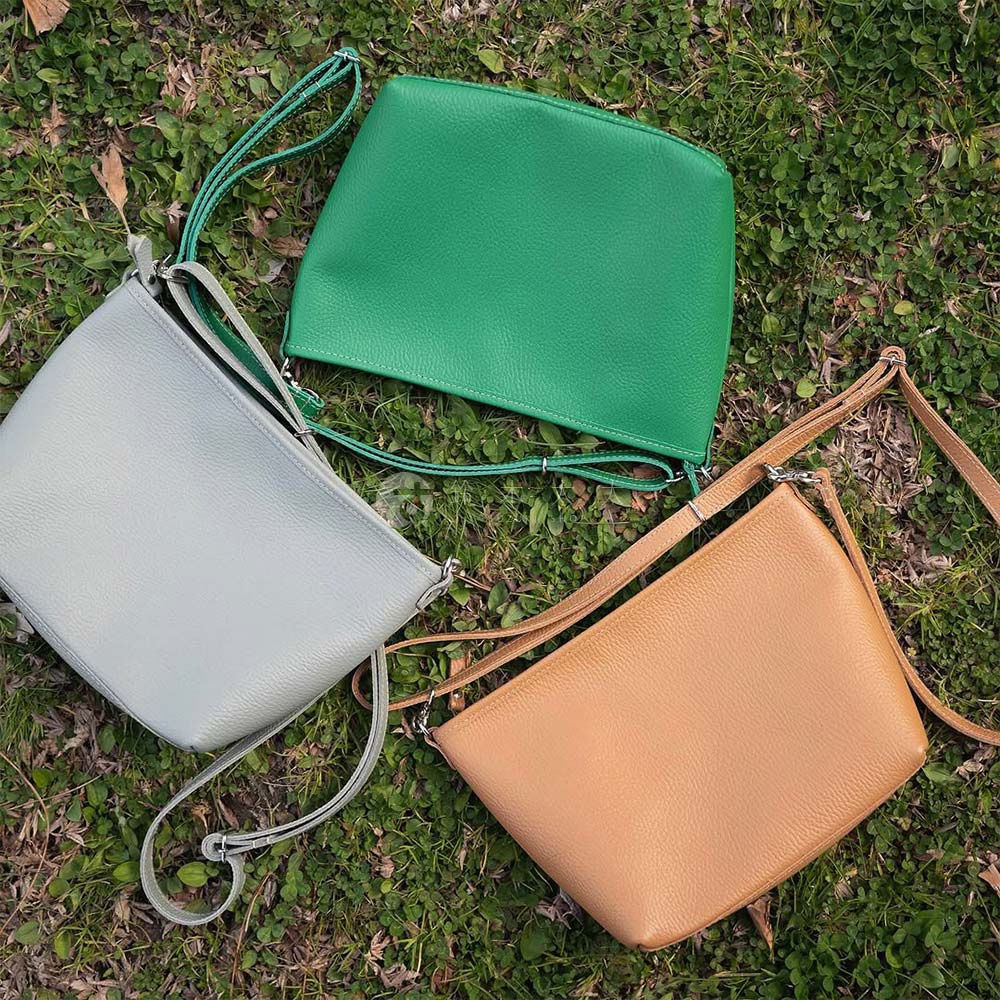 三款不同顏色（淺灰、翠綠、焦糖色）的皮革隨身側背包，皆配有細長可調式背帶，包身線條簡潔，質感紋理清晰，散落在草地上。