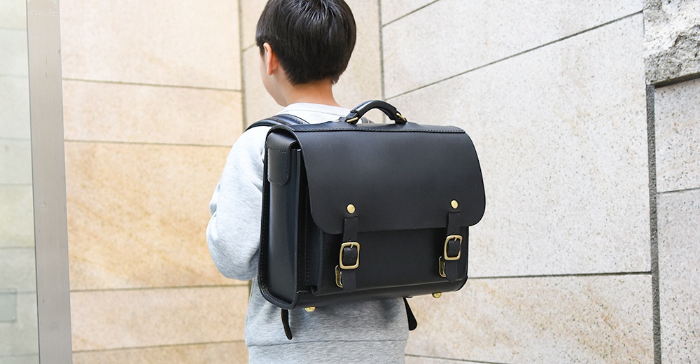 一個男孩背著一個經典黑色皮革橫式書包，書包配有金色扣環和皮帶。