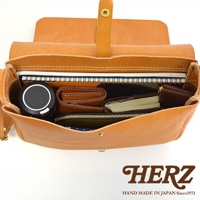 日本HERZ手工製作的棕色皮革軟皮橫式斜背包，內部裝有筆記本、錢包和圓形黑色小物，側邊有品牌標誌。