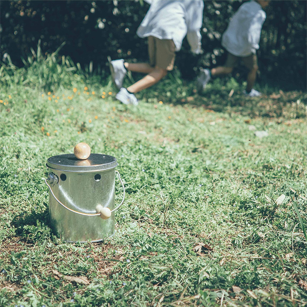 在綠草地上，一個帶有木製提手和頂蓋的鍍鋅鋼製蚊香桶，後方有兩個人影正在奔跑