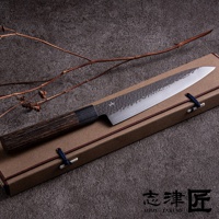 一柄21公分的不鏽鋼主廚刀，刀刃表面有槌紋，搭配深色木紋手柄，放置在米色紙盒上，盒蓋以藍色繩結固定。