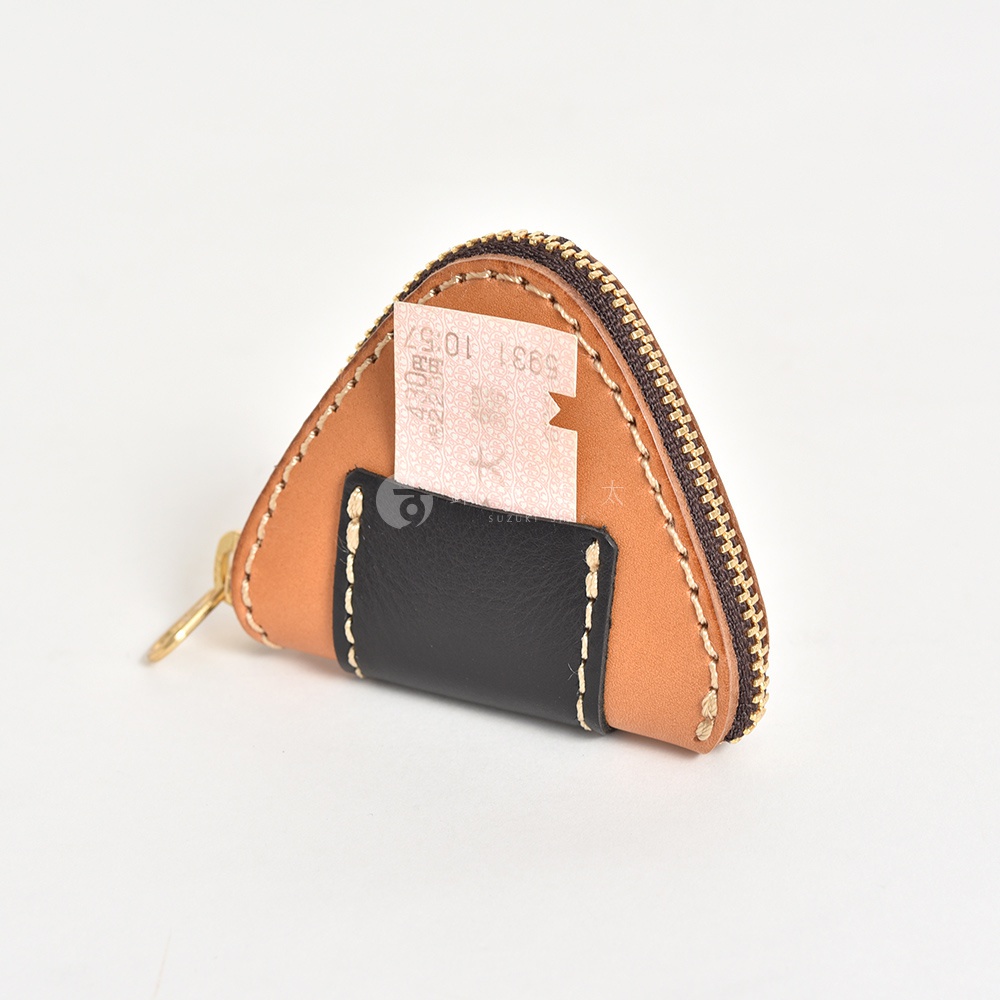 一個造型精巧的三角飯糰形狀皮革零錢包，以淺棕色皮革為主體，搭配黑色皮革拼接細節，邊緣由粗縫線固定。錢包一側縫有一個黑色皮革夾層，裡面插著一張印有日本鐵道乘車資訊的紙票。整體採用拉鍊開合設計，呈現職人手作的質感。