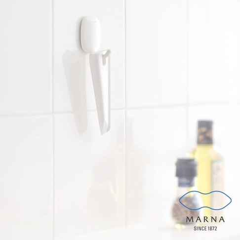 白色廚房清潔刮板，附白色掛座，掛於白色磁磚牆面上，刮板為塑膠材質，呈錐形，底部有斜切面。背景右側有兩瓶模糊的調味品。