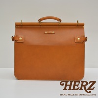 職人手作皮革旅行包 A-102，採用鞣製的淺棕色皮革製作，設有手提握把、黃銅金屬扣環和側邊扣帶，正面有一個壓印品牌標誌的皮牌，整體設計簡約大方。