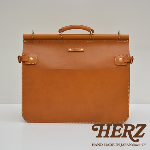 職人手作皮革旅行包 A-102，採用鞣製的淺棕色皮革製作，設有手提握把、黃銅金屬扣環和側邊扣帶，正面有一個壓印品牌標誌的皮牌，整體設計簡約大方。