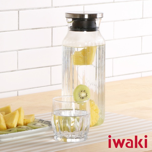 直立式耐熱玻璃水壺，帶有不鏽鋼蓋，內部裝有鳳梨塊和奇異果片。水壺側邊有垂直條紋，容量約為1公升。旁邊有一個裝有清水的玻璃杯，杯緣掛著一片奇異果。