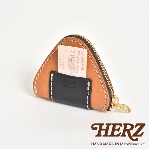 一個外型如三角飯糰的日本HERZ手工皮革零錢包，淺棕色主體搭配黑色皮革裝飾，上方插入一張印有4.28日期的鐵路票根，側面配有黃銅拉鍊與拉環，整體呈現簡約精緻的手作質感。