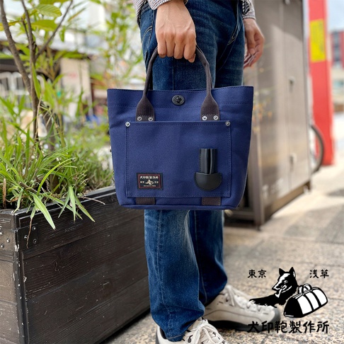 深藍色帆布製成的水桶托特包，配有深棕色提把、金屬扣飾和一個黑色皮革裝飾的前袋。包身左側有一個包含品牌標誌的織標。