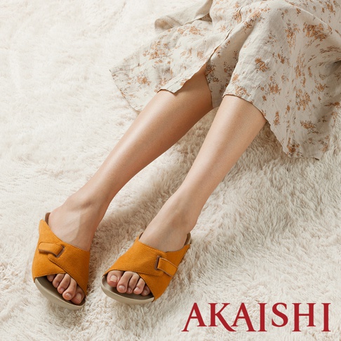 一雙黃褐色麂皮寬帶室內拖鞋，鞋面交叉設計，配有可調節的魔鬼氈，鞋底為淺卡其色。