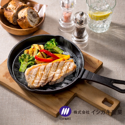 黑色鑄鐵波浪平底鍋盛有烤豬排、花椰菜、櫛瓜和彩椒，鍋子放在木製砧板上，背景有麵包、調味罐和飲品。