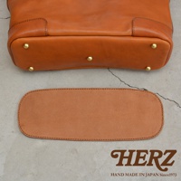 HERZ 品牌標誌，下方文字為 HAND MADE IN JAPAN Since1973，旁邊是棕色皮革托特包的底部細節，以及一個與之搭配的皮革襯墊。