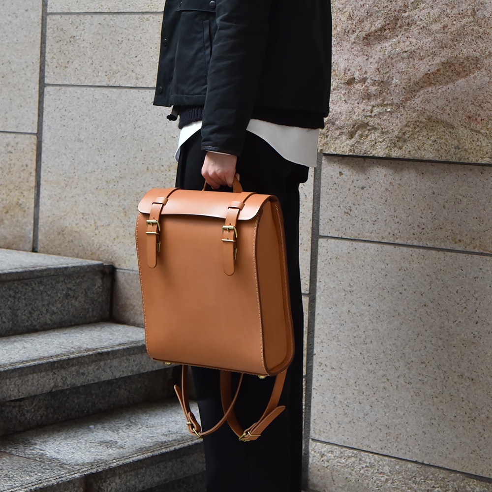 棕褐色皮革後背包，配有兩個金色搭扣和可調節背帶，由厚實皮革製成，設計簡潔。
