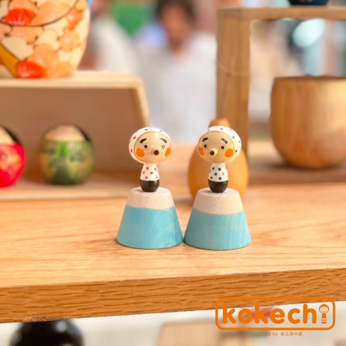 兩尊木芥子開運擺飾，身穿白色附粉紅色圓點或藍色圓點上衣，頭戴白色頭巾，臉部有圓形紅臉頰，站立於藍色山形木質底座上。