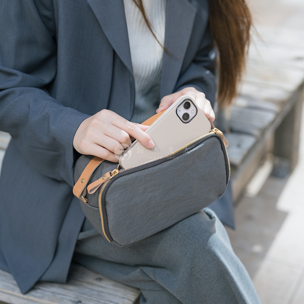 一位身穿灰色西裝外套和褲子的女性，正從一個深灰色兩用斜背包中拿出一個淺色手機。