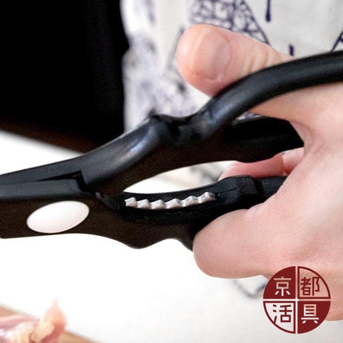 一隻手正握著一對黑色手柄的不鏽鋼萬用料理剪刀，剪刀的刀刃部分有鋸齒狀，靠近手柄處有一個圓形白色標誌。