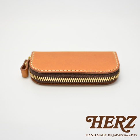 HERZ 鑰匙包，以棕褐色皮革製成，設有拉鍊開合和一個皮質拉環，底部有「HERZ HAND MADE IN JAPAN Since1973」的文字標誌。
