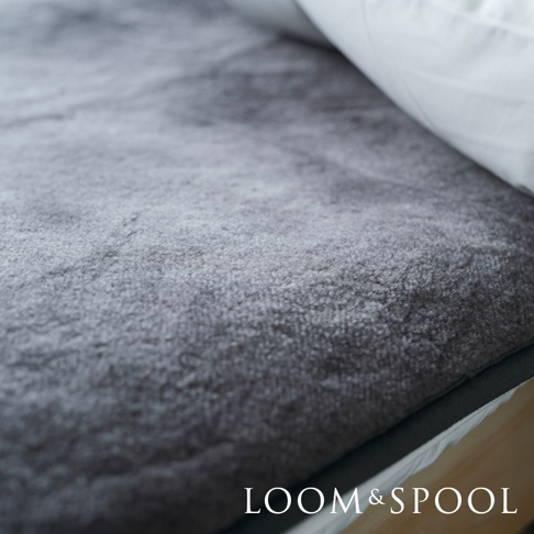 單人美麗諾羊毛保暖墊，厚實柔軟的炭灰色羊毛表面，搭配白色床單一角，展現溫暖舒適的質感。