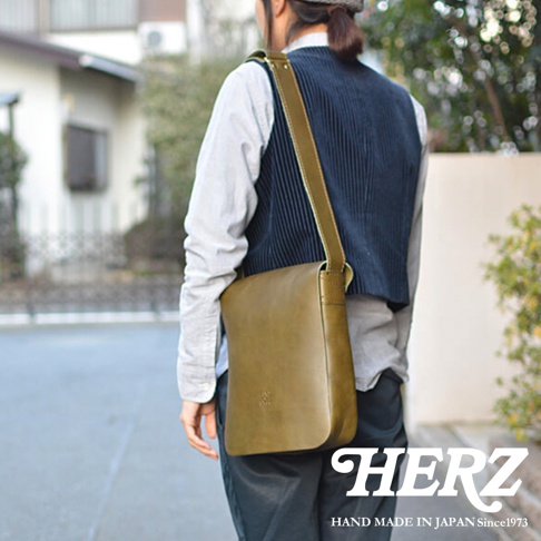 從背面拍攝，一名身穿深藍色背心和淺藍色襯衫的人，肩上挎著一個橄欖綠色的皮革斜背包，包上印有品牌標誌，下方有「HERZ HAND MADE IN JAPAN Since1973」的文字。