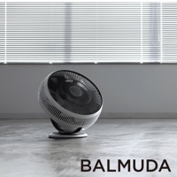 白色 BALMUDA GreenFan Cirq 循環扇，帶有黑色扇葉和網罩，放置在灰色水泥地板上，背景是帶百葉窗的白色窗戶。
