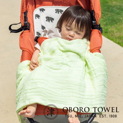 淺綠色三層紗純棉寶寶裹身毯，輕柔透氣，包裹著熟睡的嬰兒，嬰兒乘坐於橙色嬰兒車中，嬰兒車內襯印有黑色熊圖案。