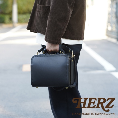 職人手作黑色皮革兩用方形單肩包，包身結構方正，配有金色拉鍊與釦環，展現簡約俐落的設計感。