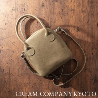 淺卡其色荔枝紋皮革手提斜背兩用包，貝殼狀包身，搭配圓潤手把及可拆卸斜背帶，置於木紋背景上。