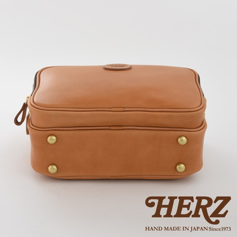 棕色皮革手作兩用公事包，方形設計，頂部有圓形皮革標誌，側邊有拉鍊和皮革拉環，底部有四個金色金屬腳釘。