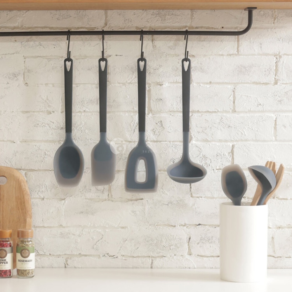櫥櫃下方，白色磚牆背景，金屬架上懸掛著數個深灰色矽膠廚具，下方右側有一個白色杯子，裝著數支深灰色及木色廚具。