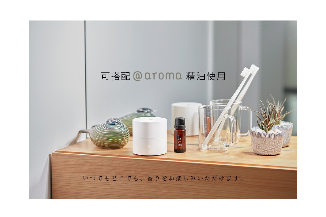 白色圓柱形隨身精油香氛機，旁有一瓶琥珀色精油，以及玻璃杯、牙刷與盆栽，擺放在木質檯面上。