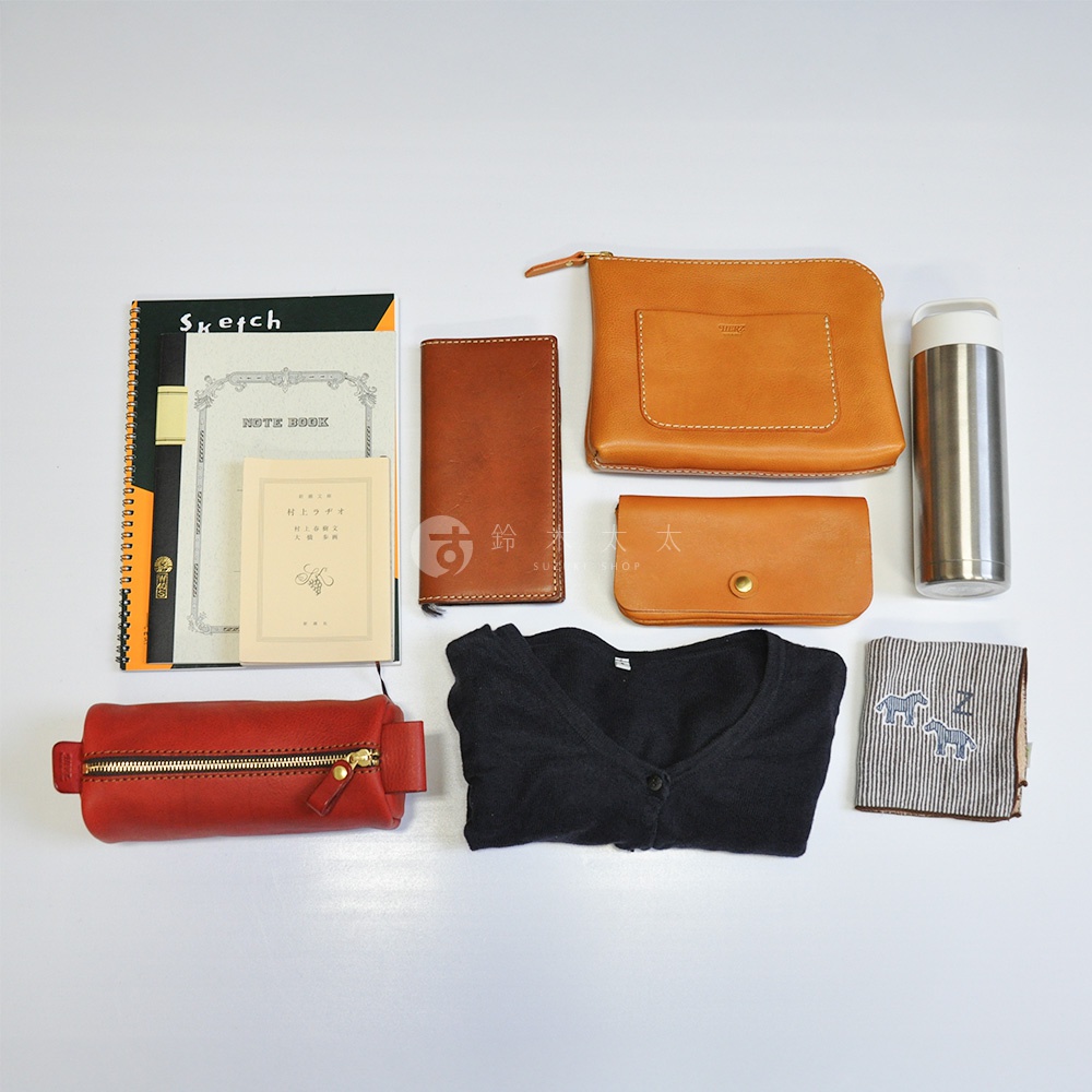 多樣皮革小物，包含筆記本、筆記本套、兩個棕色皮革夾、紅色皮革化妝包、黑色毛衣、銀色保溫瓶和條紋手帕。