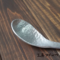 銀色鋁製湯勺，勺面佈滿錘擊紋理，置於深棕色木紋背景上。