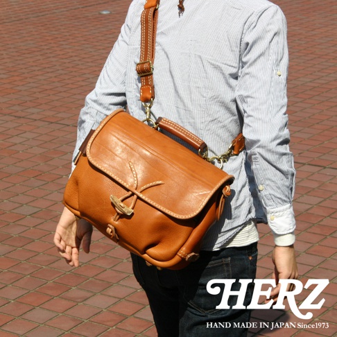 棕色皮革郵差包，採用鹿角扣和繩子設計，配有可調節皮革肩帶和提把，整體呈現職人手作質感。