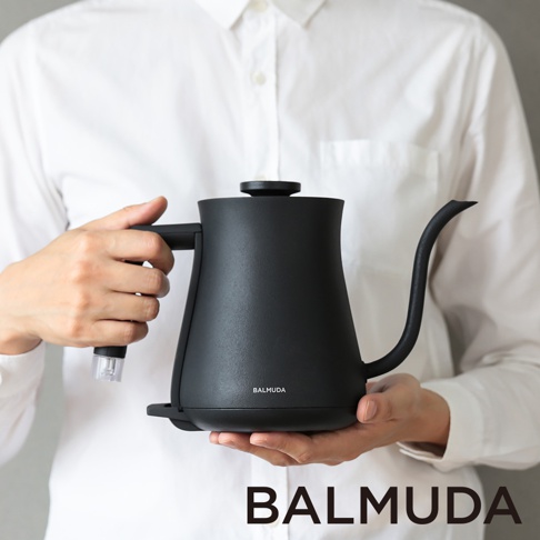 雙手捧著黑色啞光質感的 BALMUDA 絕美手沖壺，壺身呈水滴狀，配有細長彎曲的壺嘴和人體工學手把。