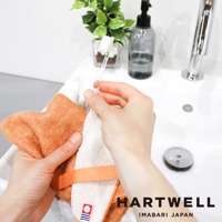 雙手正拿著一條橘色毛巾和一條白色毛巾，毛巾上印有品牌標誌。毛巾材質看起來柔軟，有紋理。旁邊有一個透明牙刷，以及一瓶黑色的皂液和一個水龍頭。